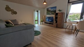 A modern living room with a grey sofa, wooden entertainment unit, and a dining area with chairs. - 2 room apartment touring view at the Kaiserberge motorcycle / bicycle garage (Waldstetten)