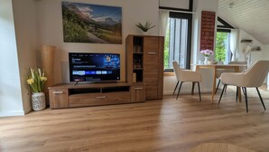 A living room with a wooden entertainment unit, a flat-screen TV, a dining table with chairs, and a large window with curtains. - 2 room apartment touring view at the Kaiserberge motorcycle / bicycle garage (Waldstetten)