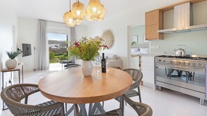 A modern kitchen with a round wooden table, wicker chairs, a stainless steel stove, and a large window offering a view of the outdoors.