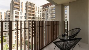 Een balkon met uitzicht op hoge wooneenheden, een glazen tafel en twee zwarte stoelen.