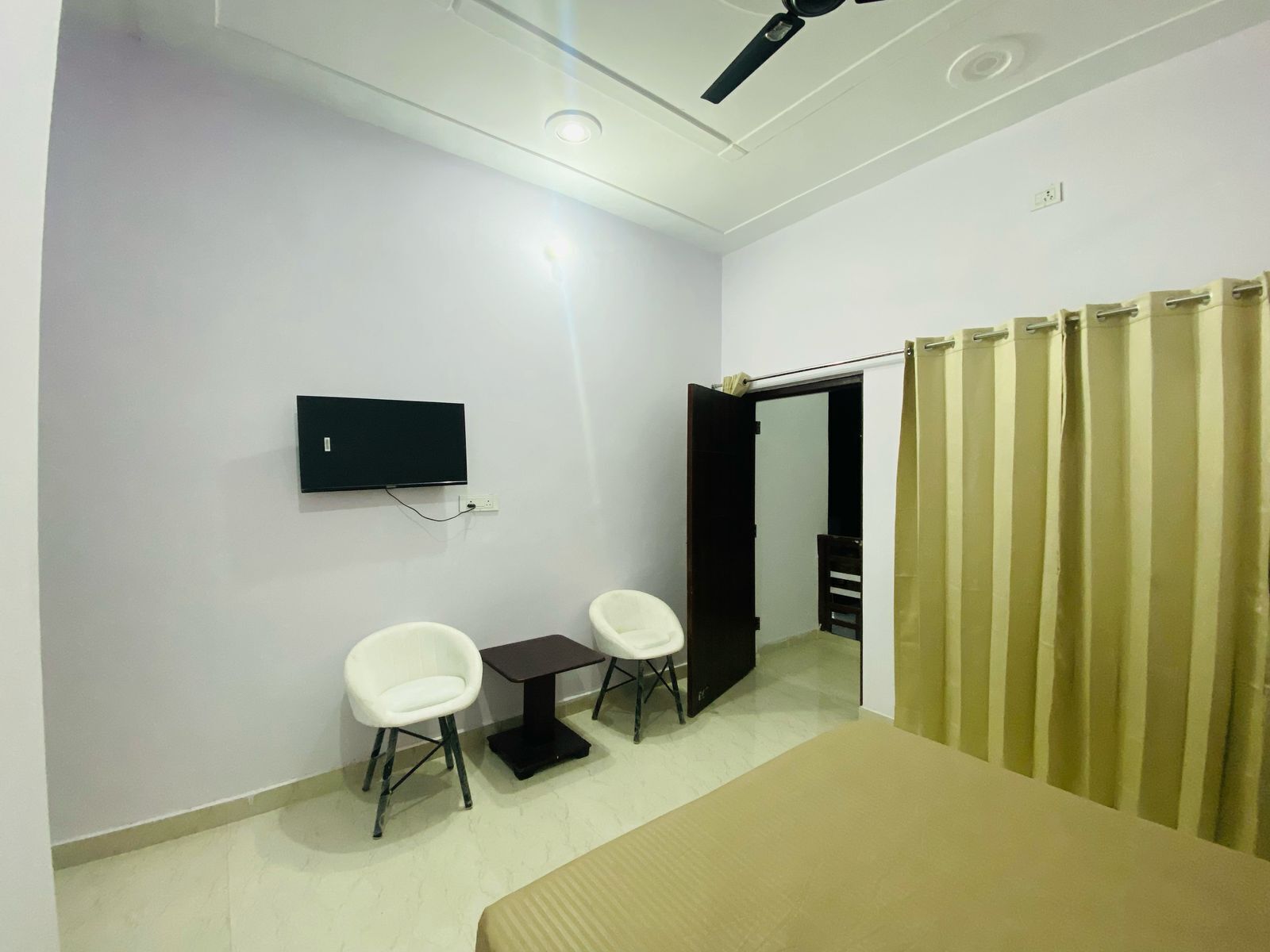 Una habitación con cama, televisor montado en la pared, dos sillas, una mesita y una puerta que da a otra estancia.