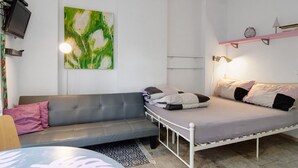 Een kamer met een grijze bank, een bed met kussens, een wandtelevisie, een schilderij en een plank met boeken en een vaas.