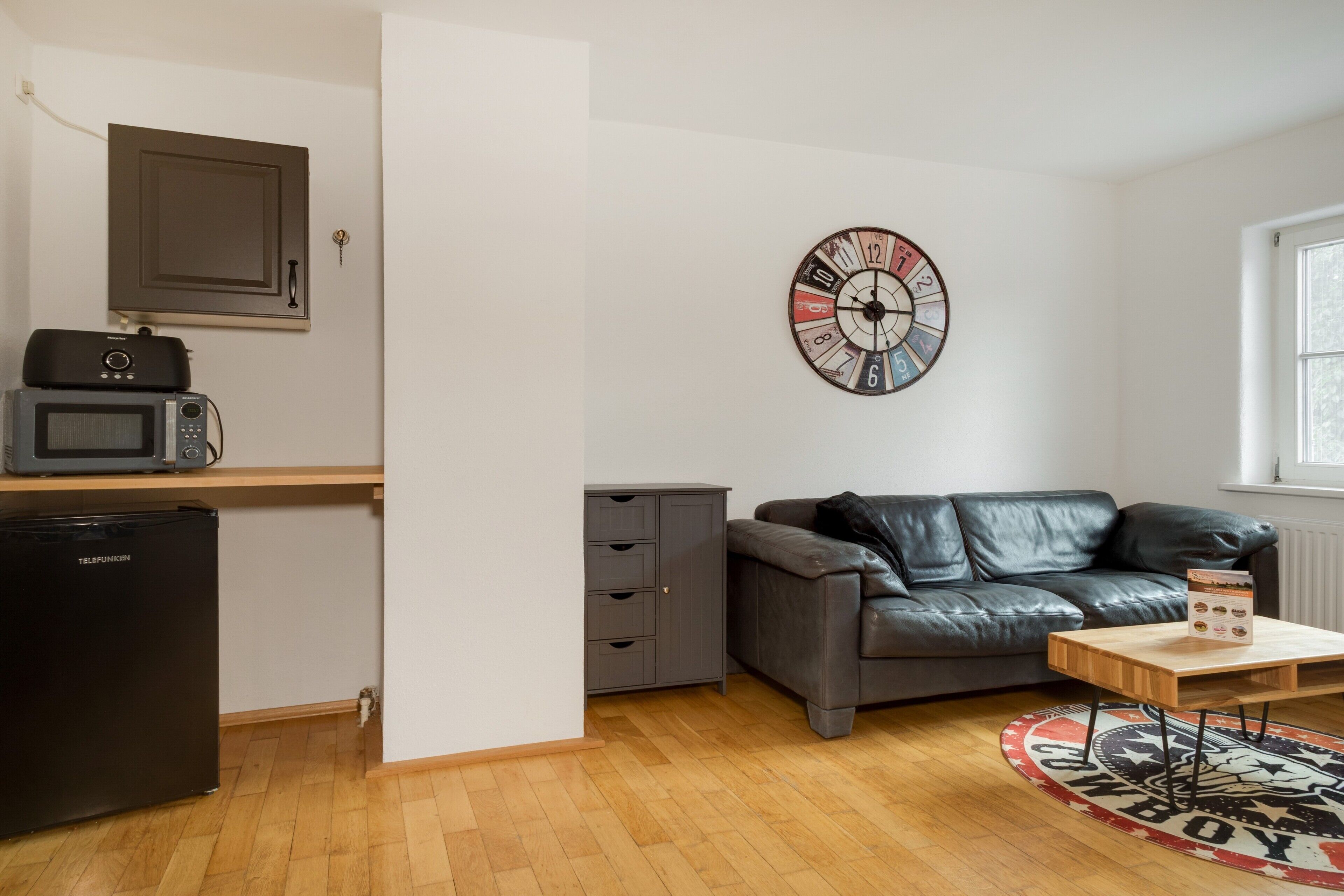 Un espace de vie compact comprenant un coin cuisine, un canapé en cuir, une horloge murale et une petite table.