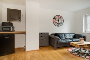 A compact living space with a kitchenette, a leather sofa, a wall clock, and a small table. - Apartment 'Mit Giebelzimmer' with Shared Garden and Wi-Fi (Ravensburg)