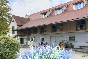Ein zweigeschossiges Gebäude mit rotem Ziegeldach, zahlreichen Fenstern und einer überdachten Terrasse mit einer Bank.