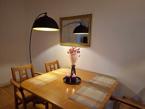 Une table à manger avec des chaises, un vase contenant des fleurs séchées, une bougie, un set de table, un miroir avec un cadre doré et une lampe sur pied.