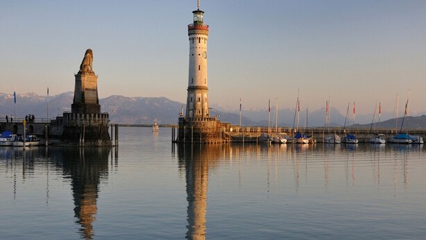 Un puerto tranquilo con dos faros destacados y una estatua, rodeado de barcos y montañas al fondo.