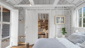 Une chambre avec un grand lit, un plancher en bois, une porte blanche donnant sur une salle de bain et une vue sur un jardin par une fenêtre.