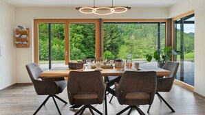 A modern dining area with a wooden table, four chairs, and a pendant light. Large windows offer a view of a forested landscape.
