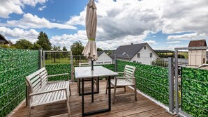 Een houten terras met een tafel, stoelen, een overdekte parasol en een metalen hek.