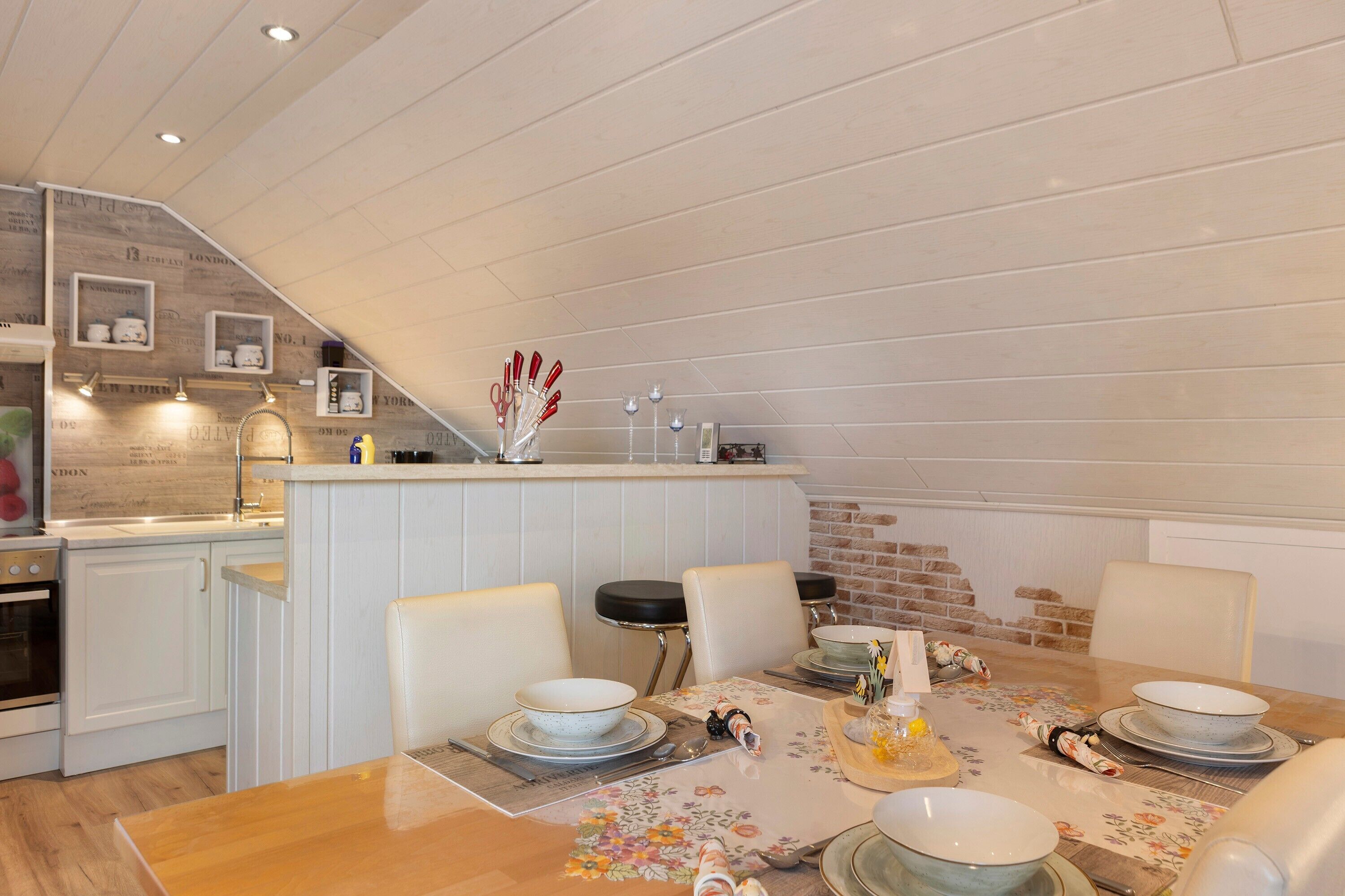 Un coin repas avec une table en bois dressée pour quatre personnes, une cuisine avec des meubles blancs et un mur en briques.