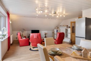 Un coin repas avec une table dressée pour deux personnes, une chaise rouge, une chaise blanche et un téléviseur en arrière-plan.