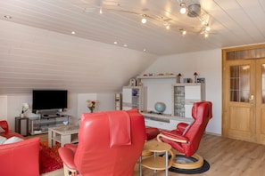 Ein modernes Wohnzimmer mit einem Flachbildfernseher, roten Ledersesseln, einem kleinen Tisch und einer Holztür.