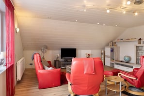 Ein Wohnzimmer mit Fernseher, roten Ledersesseln, einem kleinen Tisch und einem Bücherregal.
