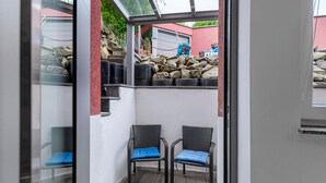A covered outdoor seating area with two chairs and a small table, a stone wall, and a view of a building in the background. - Apartment 'In Der Vulkan-Eifel' with Wi-Fi (Mendig)