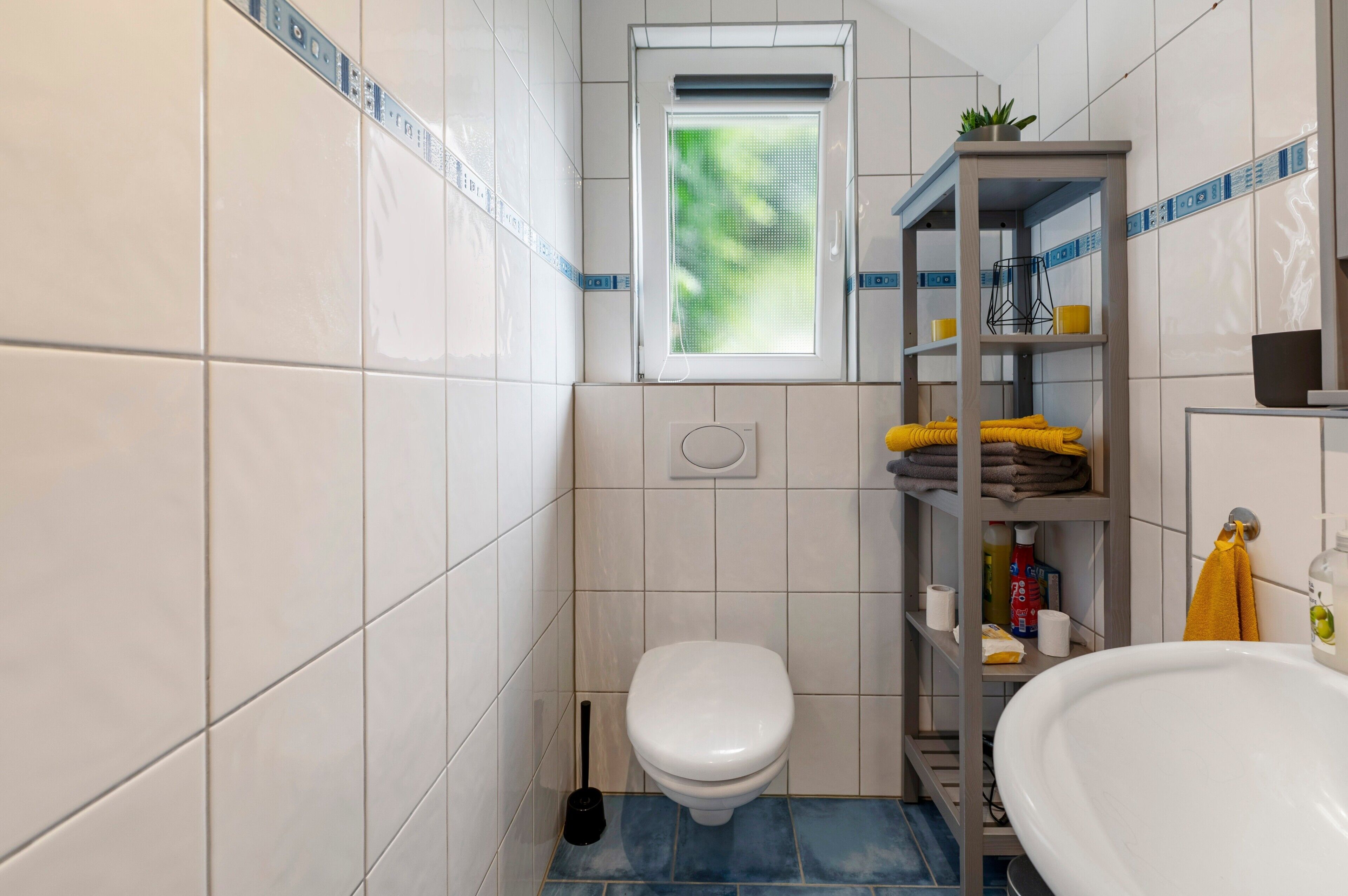 A small bathroom with a toilet, a shelf with towels, a window, and a sink.