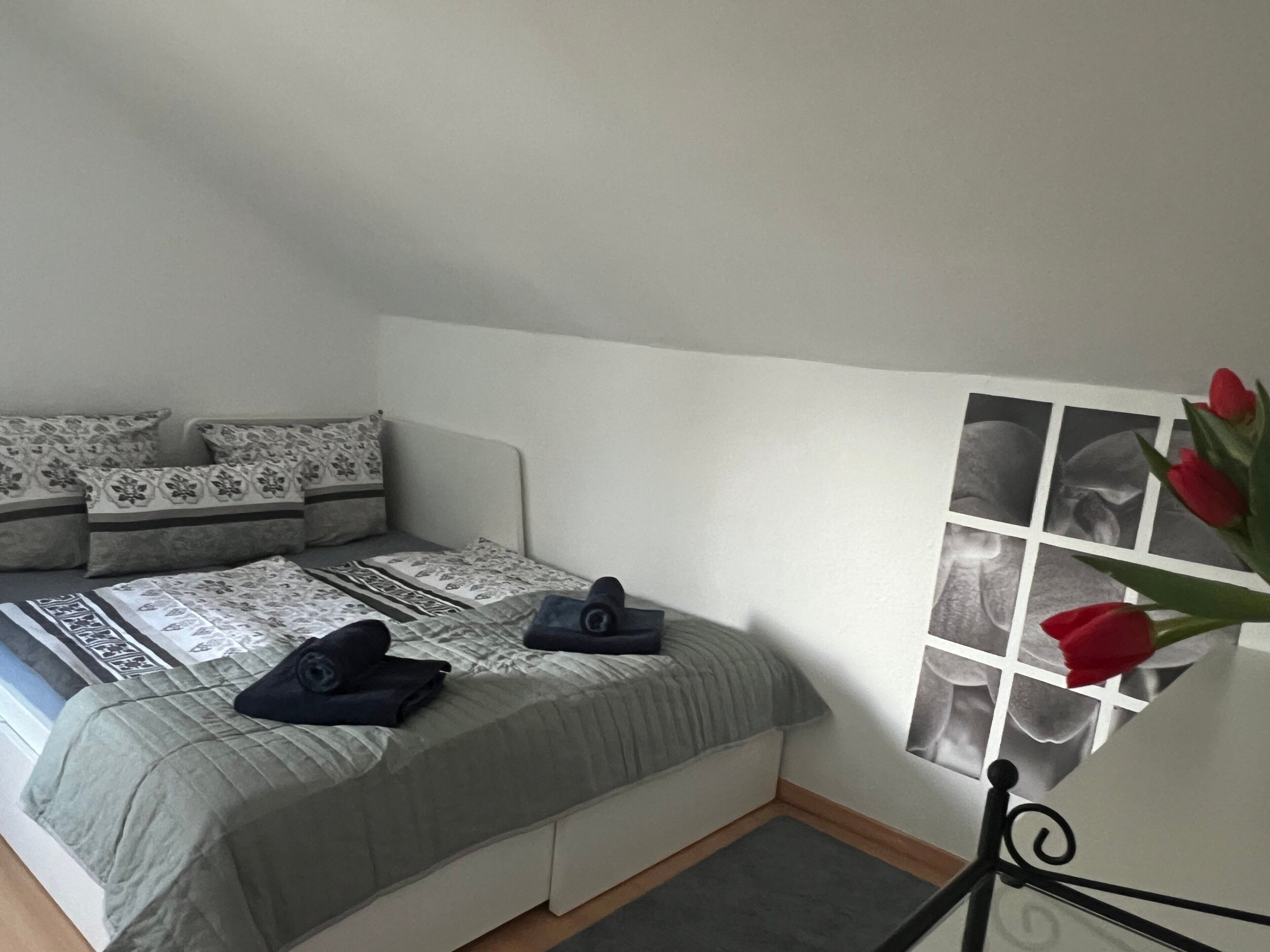 Une chambre à coucher avec un lit, des oreillers, un petit tapis et un cadre photo fixé au mur, dans lequel est disposée une composition florale.