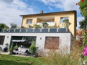 Una casa moderna de dos pisos con un balcón, un espacio de estar al aire libre cubierto y un jardín.