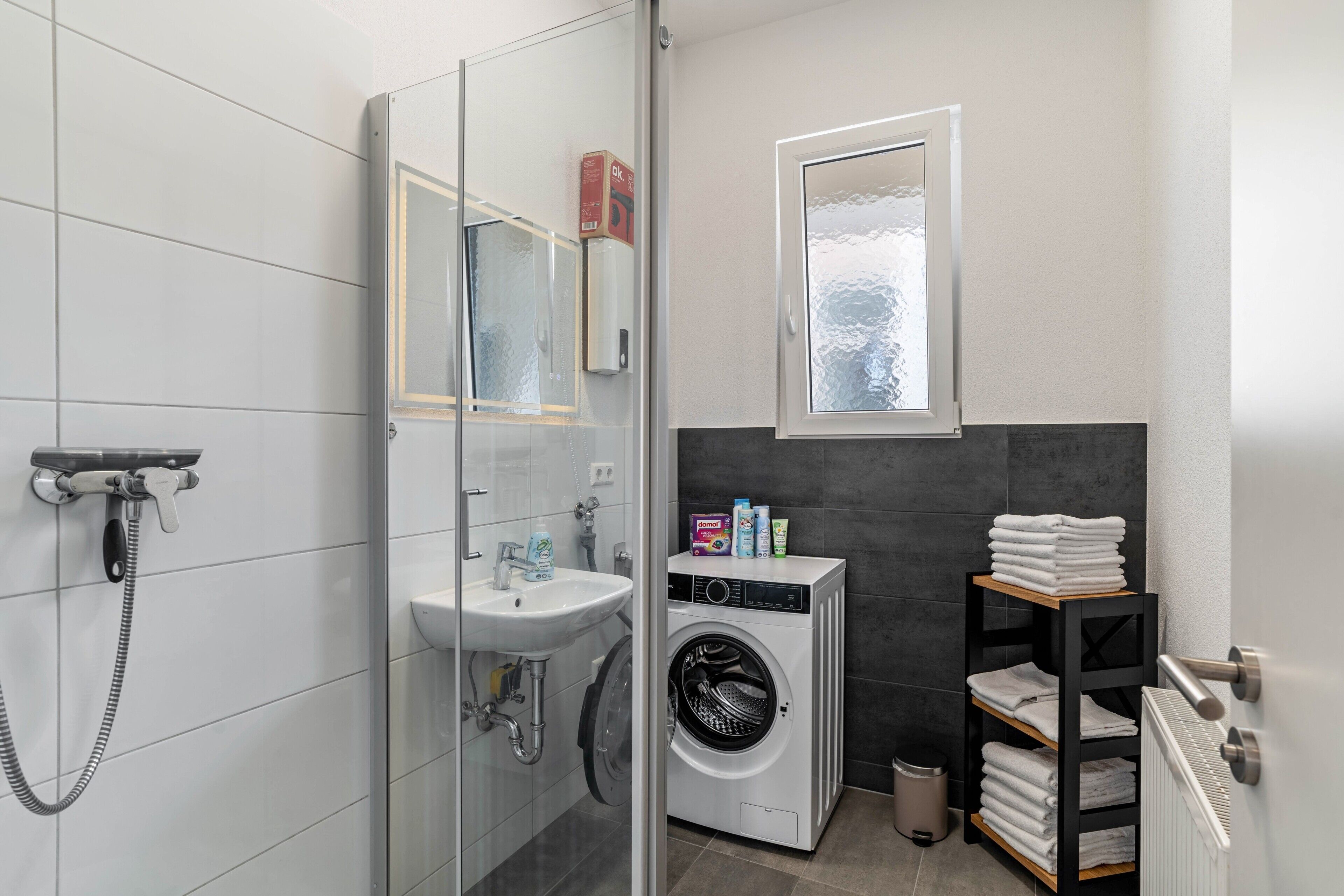 A modern bathroom with a glass shower enclosure, a built-in sink, a washing machine, and a towel rack.