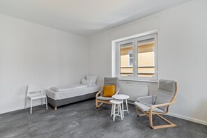 A room with a bed, a white chair, a grey lounge chair, a small white table, and a window with blinds.