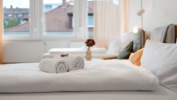 A neatly made bed with rolled white towels, a vase with flowers, and a view of buildings through a window. - Apartment '2nd Floor Left' with Wi-Fi (Pforzheim)