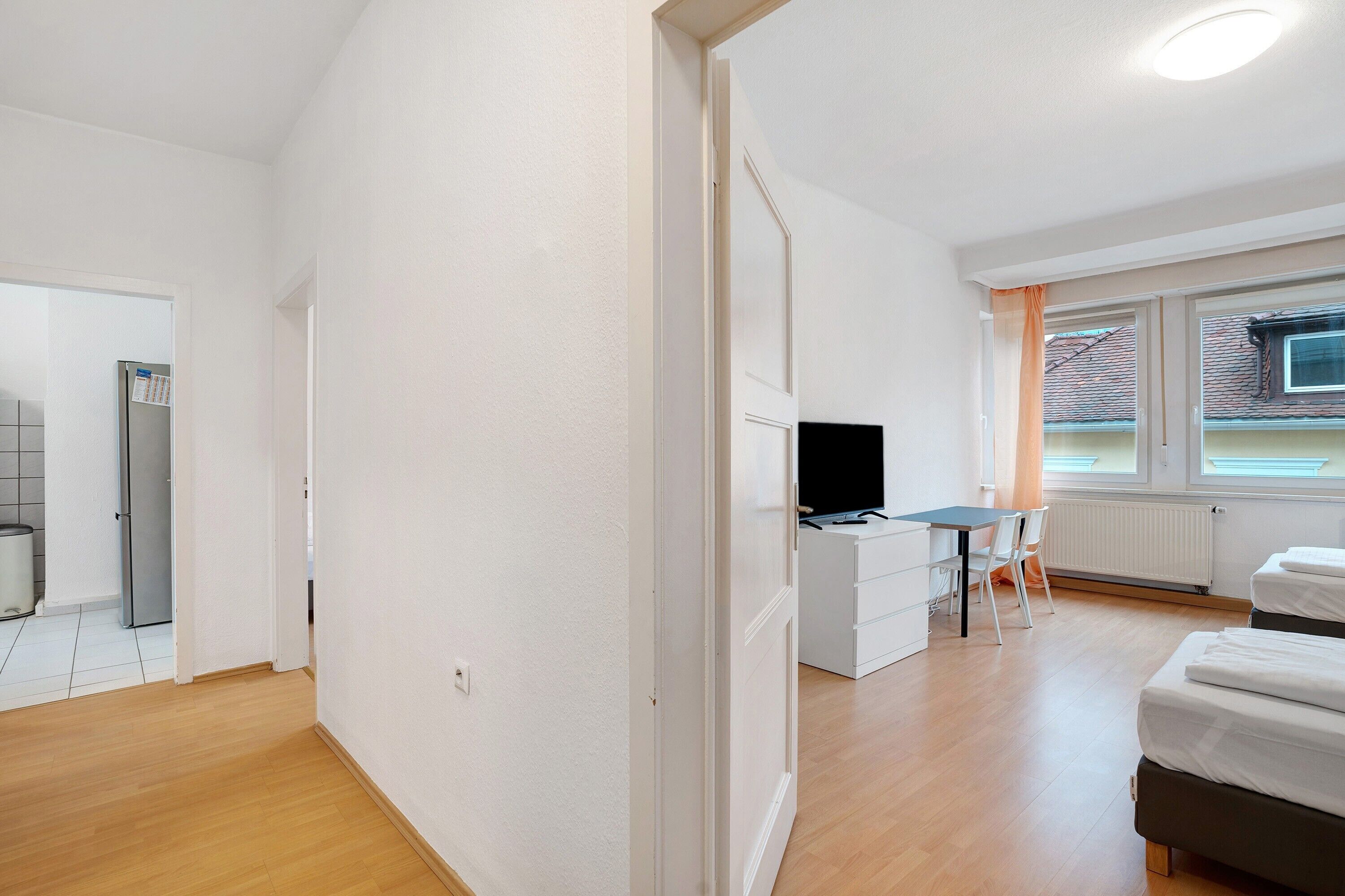 Ein moderner, gut beleuchteter Raum mit Holzboden, einem Schreibtisch mit Computer, einem Bett und einem Fenster mit Jalousien.