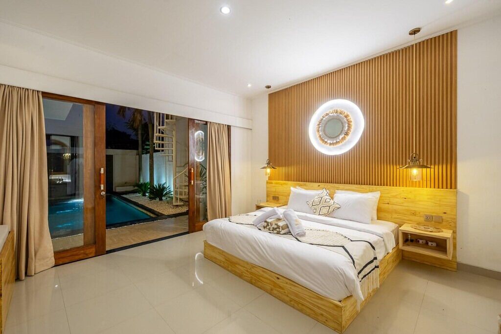 A bedroom with a wooden headboard, a large bed with white bedding, two bedside tables, and a circular mirror on the wall.