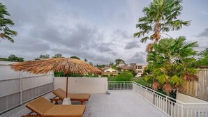 A rooftop terrace with lounge chairs and a thatched umbrella, surrounded by palm trees and a view of residential buildings. -  Umber Rise Villa (Bali)