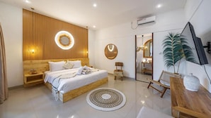 A modern bedroom with a wooden headboard, a round wall light, a bed with white linens, a small side table, a mirror, a plant, and a wooden bench. -  Umber Rise Villa (Bali)