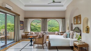 Une chambre spacieuse avec un grand lit, un coin salon comprenant un canapé et des fauteuils, une petite table et une vue sur une végétation luxuriante grâce à de grandes fenêtres.