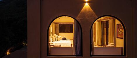 A room with two arched windows, a bed with pillows, a nightstand, and a wall-mounted light fixture.