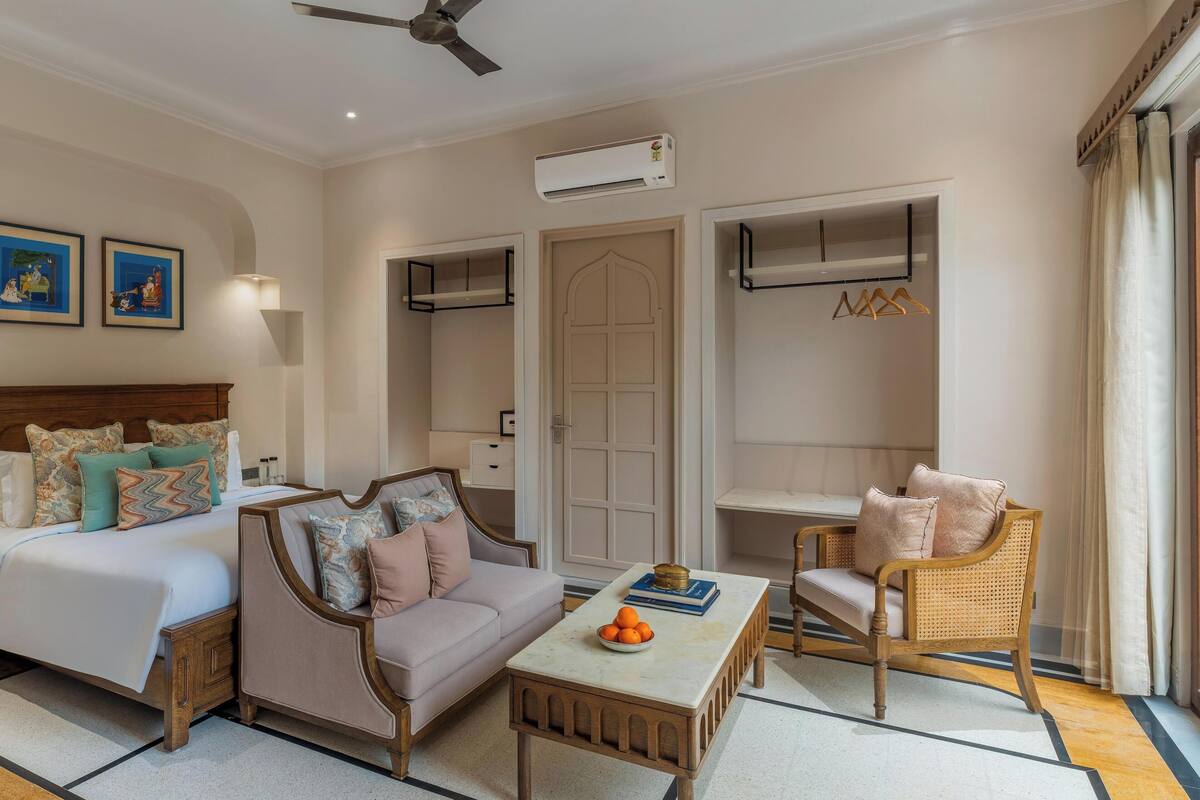 Una habitación de hotel con una cama, un sofá, una silla, una pequeña mesa con naranjas y un equipo de aire acondicionado.