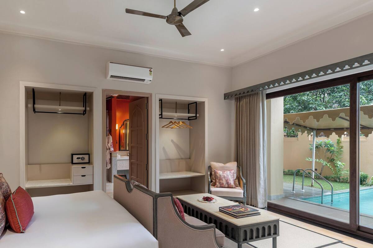 Habitación de hotel con cama, zona de estar, mesa de comedor, ventilador de techo, aire acondicionado y vistas a una piscina.