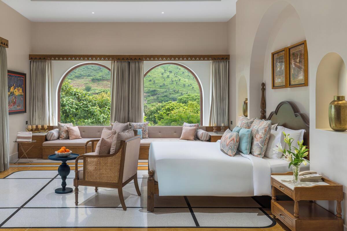 Un dormitorio con una cama grande, un sofá, una silla, una mesita con un frutero, y vistas a zonas verdes a través de ventanas arqueadas.