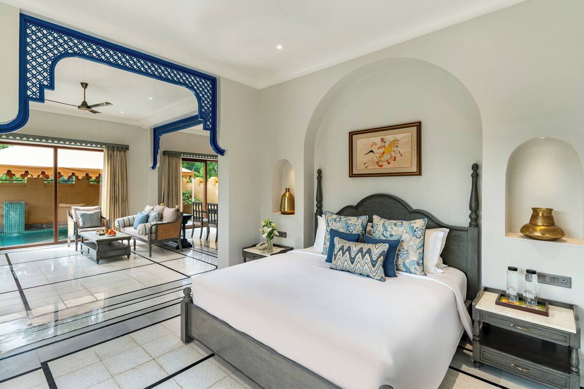 Habitación de hotel con una cama grande, zona de estar con sofá y sillones, una mesita y un balcón con piscina.