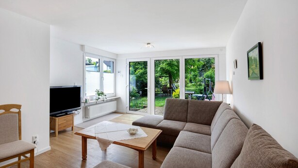 A living room with a sofa, a television on a stand, a table with a placemat, and a view of a garden through large windows.