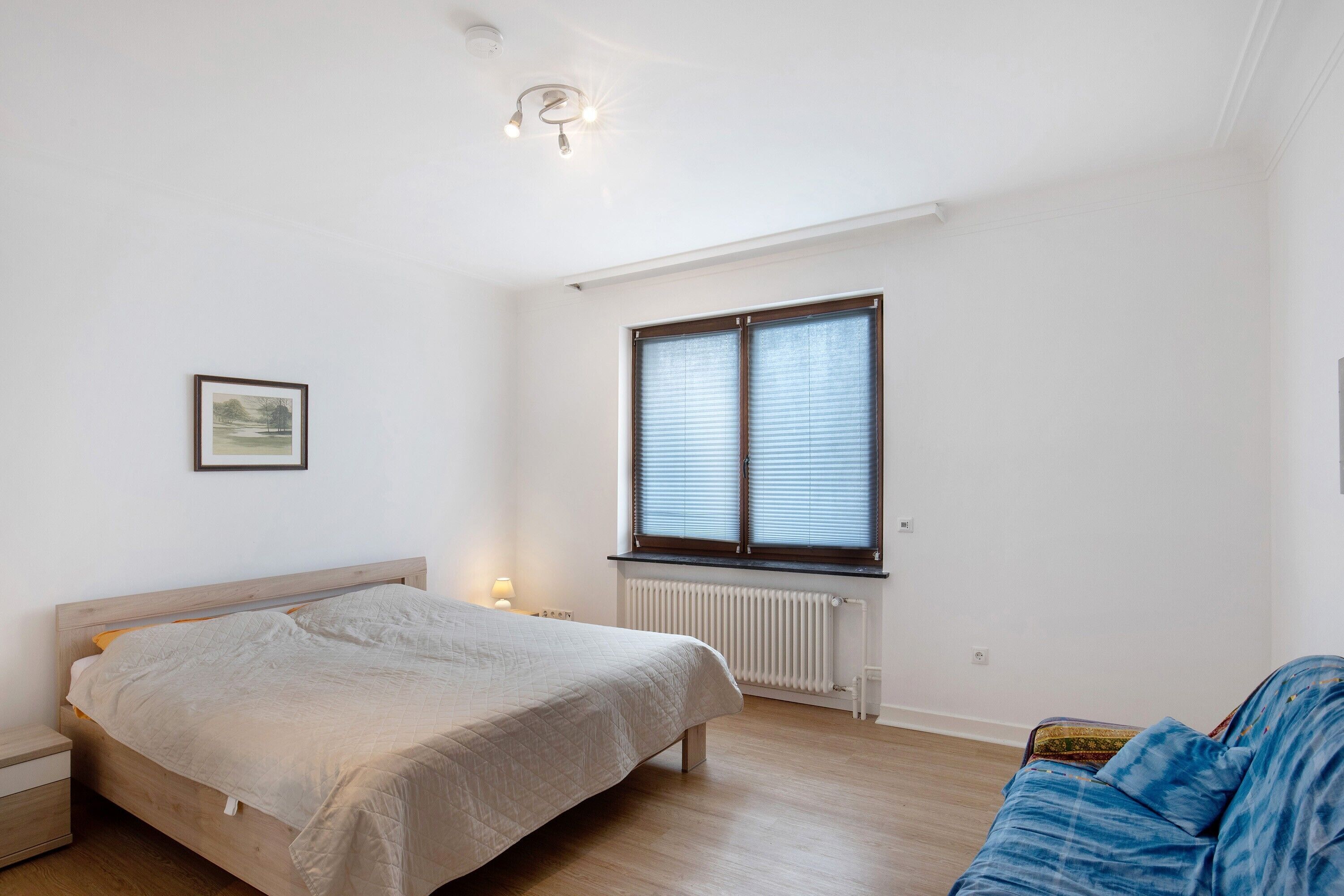 Een slaapkamer met een bed, een raam met zonwering, een radiator, een nachtkastje en een bank.