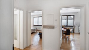 A hallway leading to a bedroom with a bed, a dining area with a table and chairs, and a kitchen area with a window.