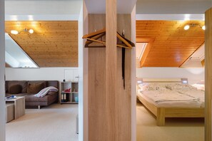 A modern bedroom with a sloped ceiling, wooden beams, a bed with floral bedding, and a small seating area with a sofa and side table.