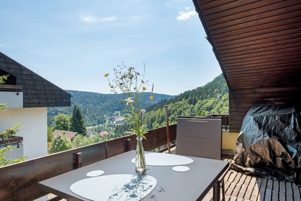A balcony with a table, a vase with flowers, and a view of a forested valley.