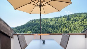 Ein Balkon mit Tisch und Stühlen, einem Sonnenschirm und Blick auf einen bewaldeten Hang.