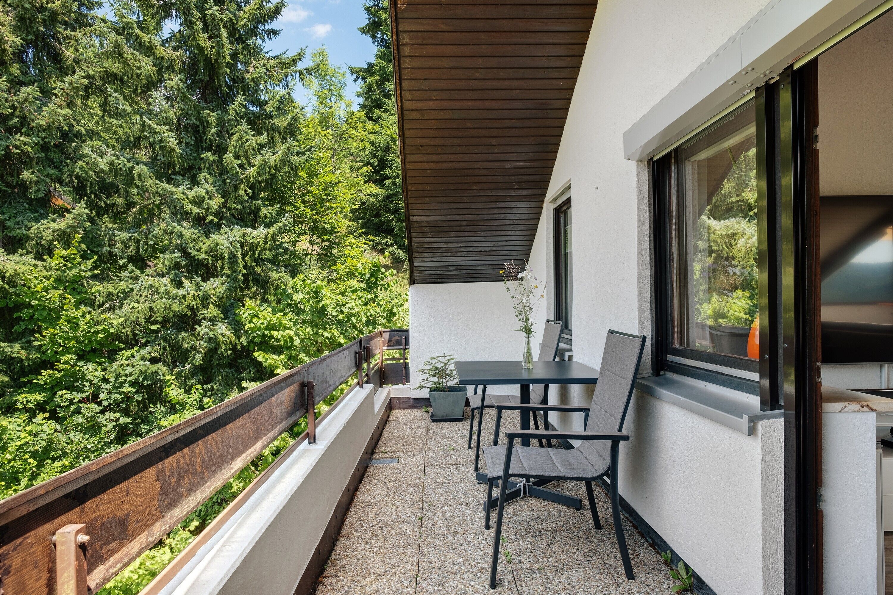 A balcony with a table and chairs, a wooden railing, and a view of dense greenery.