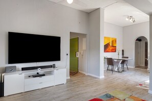 A modern living room with a large flat-screen TV, a white entertainment unit, a dining area with a table and chairs, and a colorful rug.
