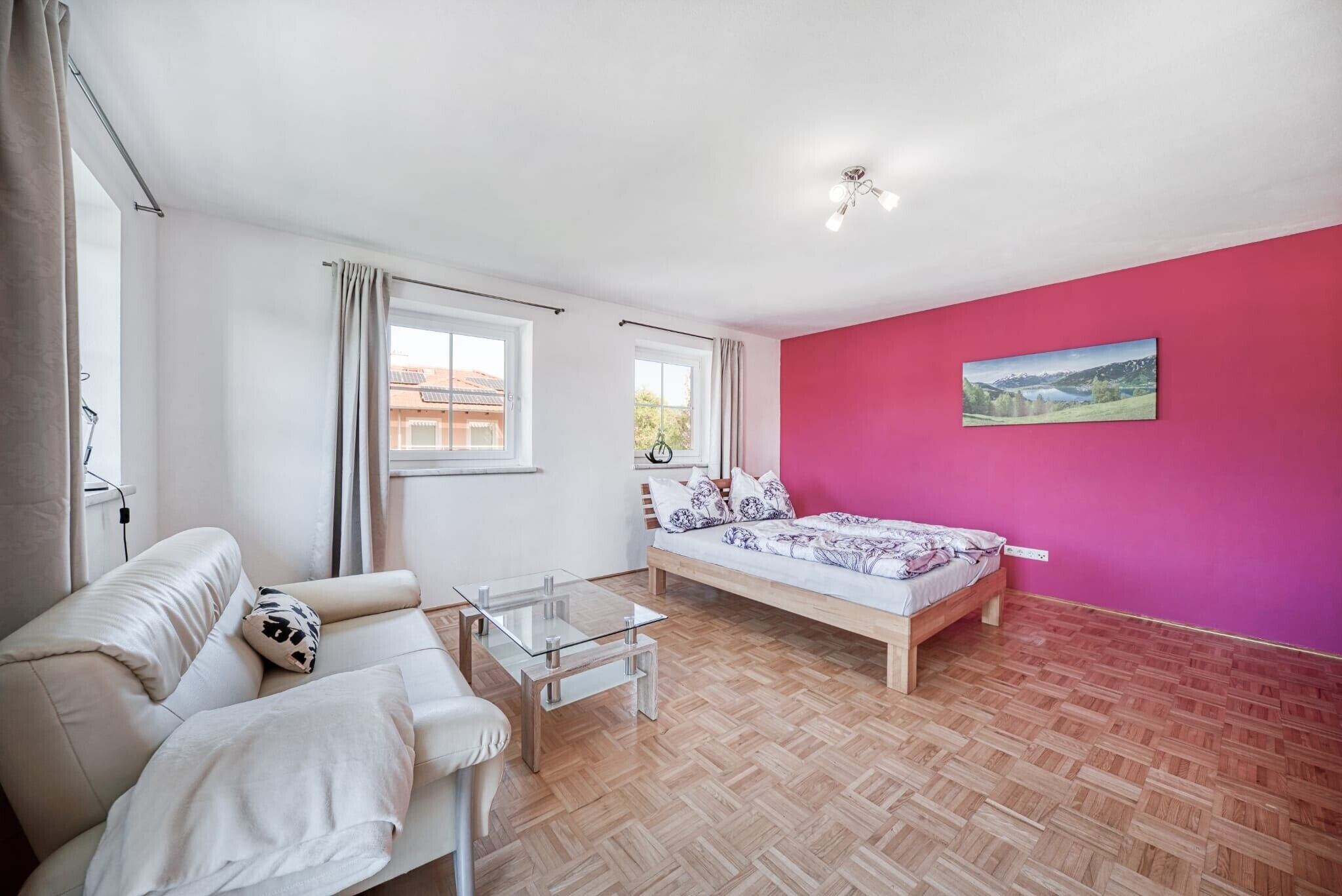 A bedroom with a bed, a sofa, a glass coffee table, a window, and a pink wall.