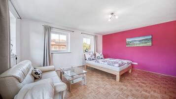 A bedroom with a bed, a sofa, a glass coffee table, a window, and a pink wall.