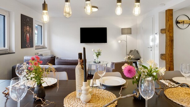 Un comedor con una mesa dispuesta para dos personas, un televisor montado en la pared y una lámpara moderna sobre la mesa.