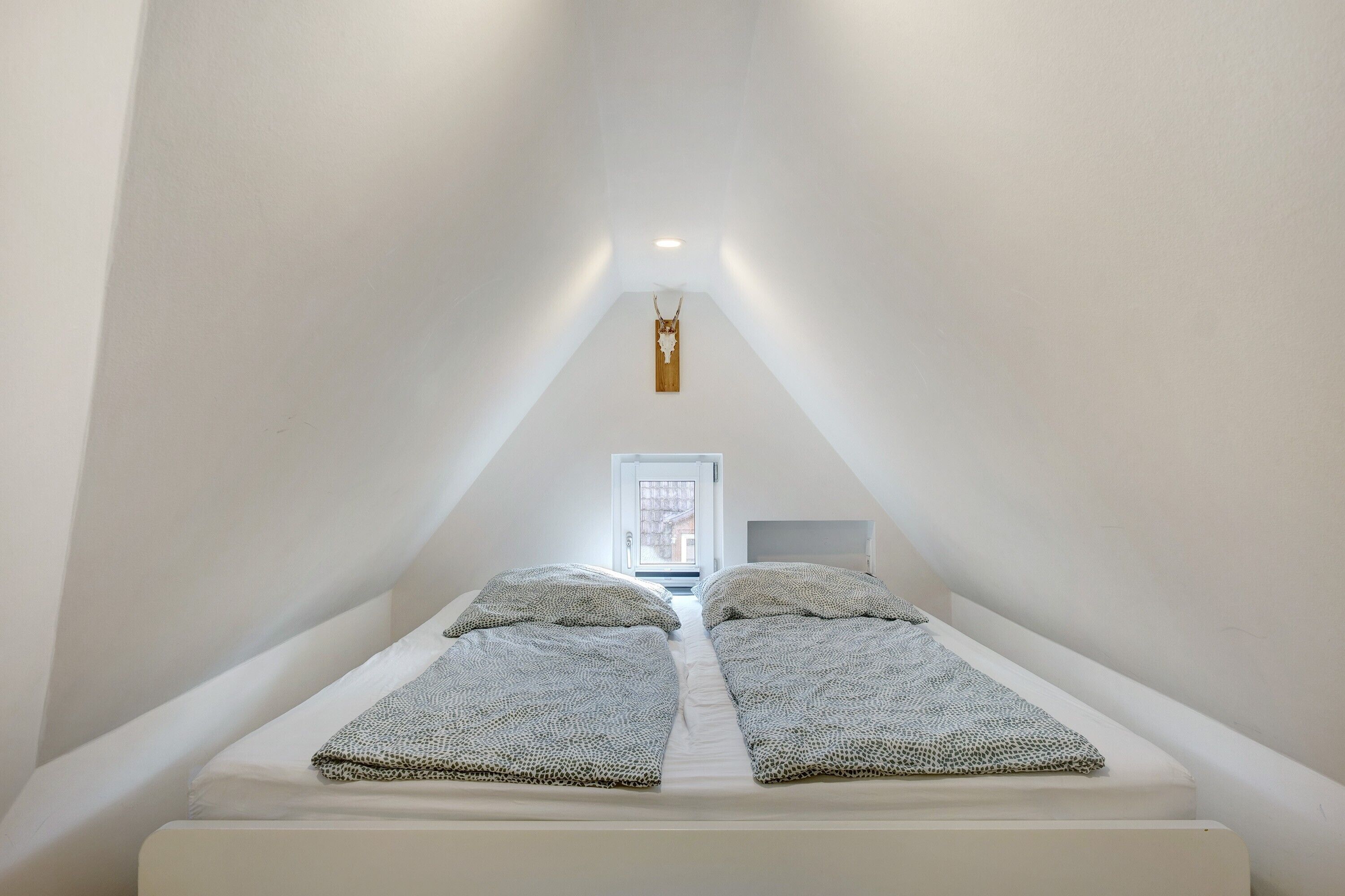 Un dormitorio bajo cubierta, estrecho, con dos camas, cabecero de madera y un ventanal pequeño.