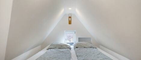 Un dormitorio bajo cubierta, estrecho, con dos camas, cabecero de madera y un ventanal pequeño.