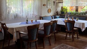 Uma sala de jantar com mesas e cadeiras de madeira, cortinas de renda e vista para o verde lá fora.