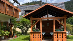 Um gazebo de madeira com telhado inclinado, cercado por um gramado bem cuidado e vasos de plantas.